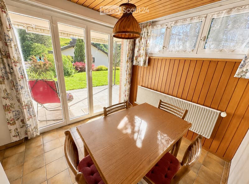 Dining room with wooden table, red upholstered chairs, radiator, and large windows with curtains. A red lounge chair is visible outside the sliding glass doors.