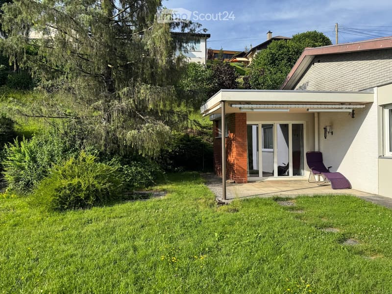 A well-maintained garden with a lawn, a patio area, and a lounge chair near the house entrance.