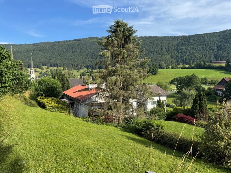 A picturesque rural landscape with a house, lush green fields, and a church in the distance, surrounded by trees and hills.