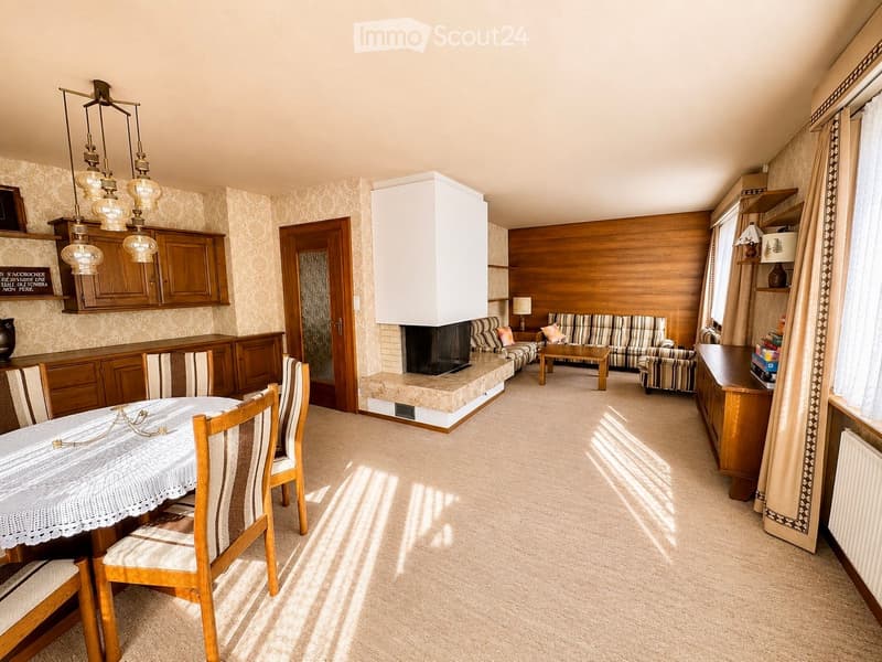 A well-furnished room with a dining area, living area, and fireplace. Features include a patterned sofa, coffee table, dining table with chairs, wooden cabinets, and a chandelier.