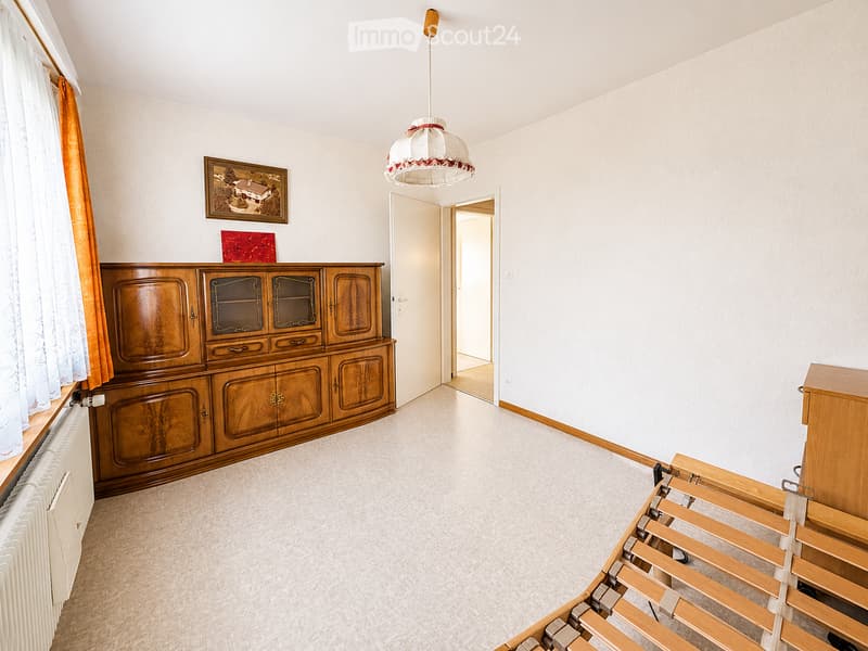 A furnished room with a large wooden cabinet, a picture frame on the wall, a radiator, and a wooden bed frame visible in the foreground.