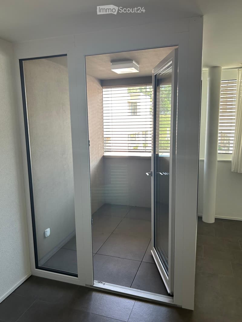 A modern hallway with a large glass sliding door leading to a balcony area, featuring tiled flooring and white walls.