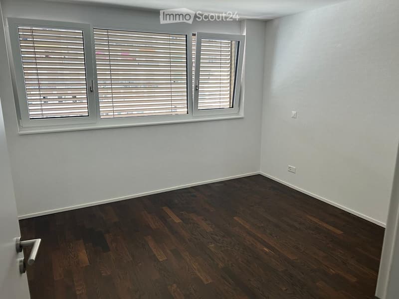 Empty room with light grey walls, dark wooden flooring, and three windows with white blinds. No furniture or equipment is present.