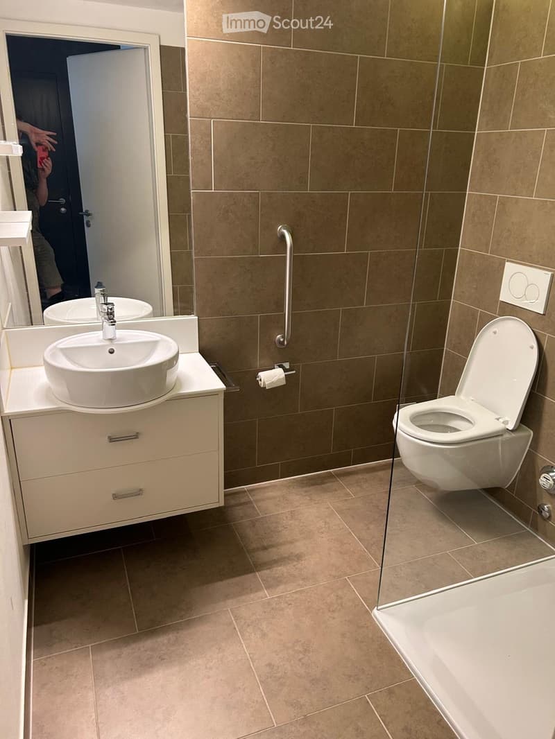 Modern bathroom with a white sink, faucet, toilet, and a glass-enclosed shower area. Beige tiles on walls and floor, with a bathroom cabinet beneath the sink.