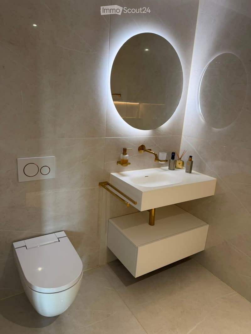 Modern bathroom with a white sink, gold faucet, round mirror, and a toilet. The sink has a built-in cabinet and a towel rack below.
