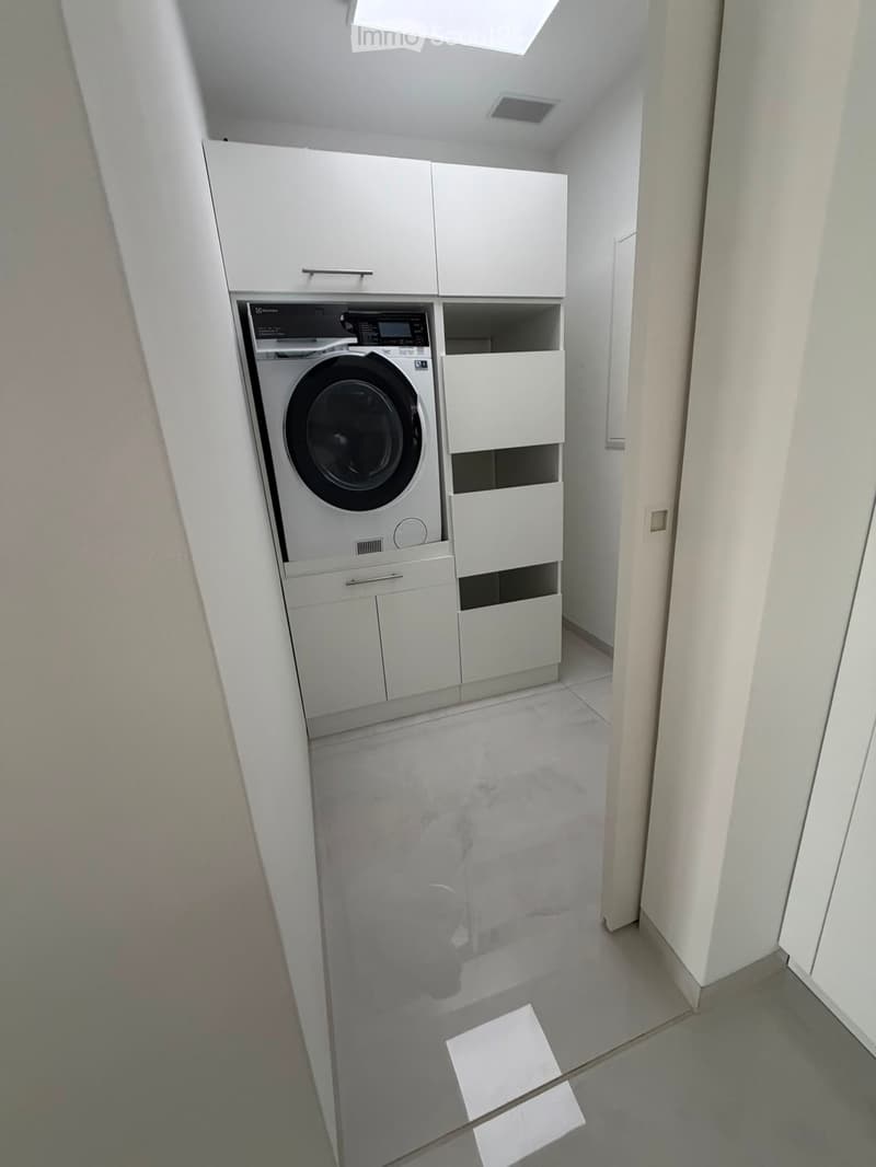 A narrow hallway with a white washing machine and matching cabinets, light grey tiled floor, and white walls.