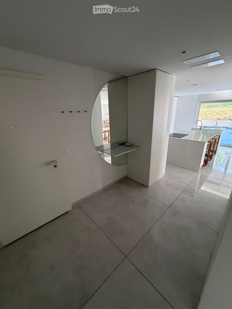 Empty hallway with tiled floor, white walls, and a curved mirror on the wall. The hallway leads to a modern kitchen area visible in the background.