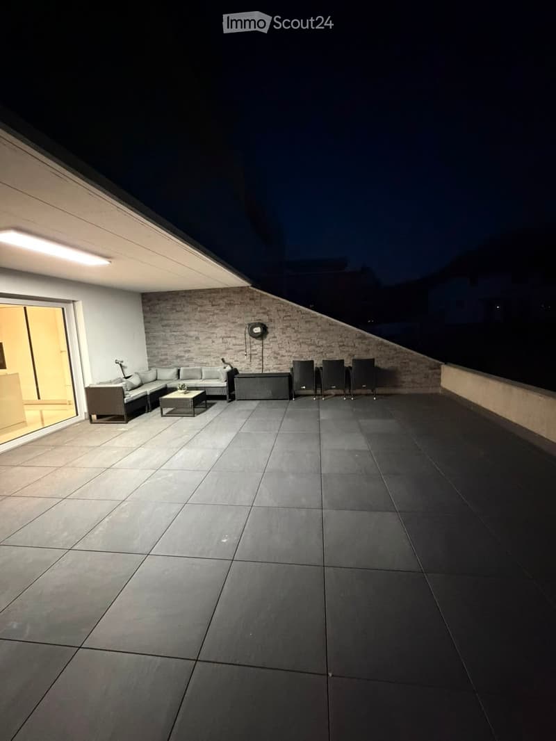 Balcony with grey tiled floor, sectional sofa, coffee table, and chairs arranged against a brick wall.
