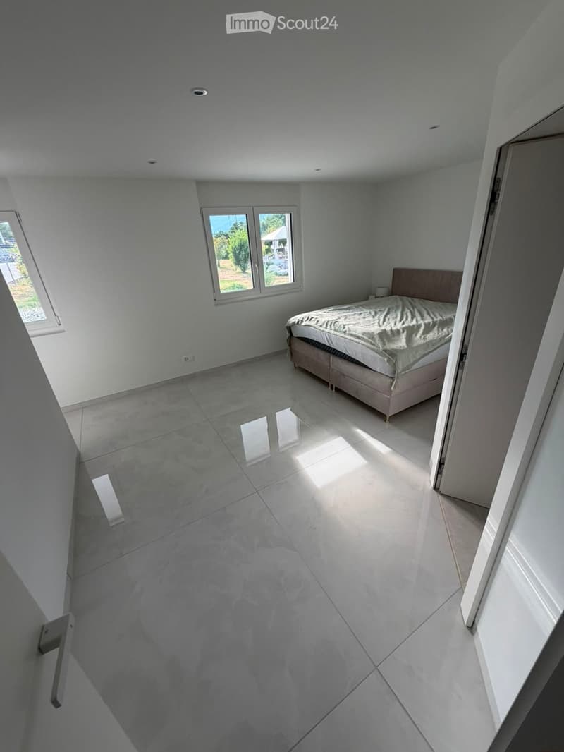 Modern bedroom with a large bed, white walls, and large windows allowing natural light to fill the space. The floor is made of polished concrete tiles.
