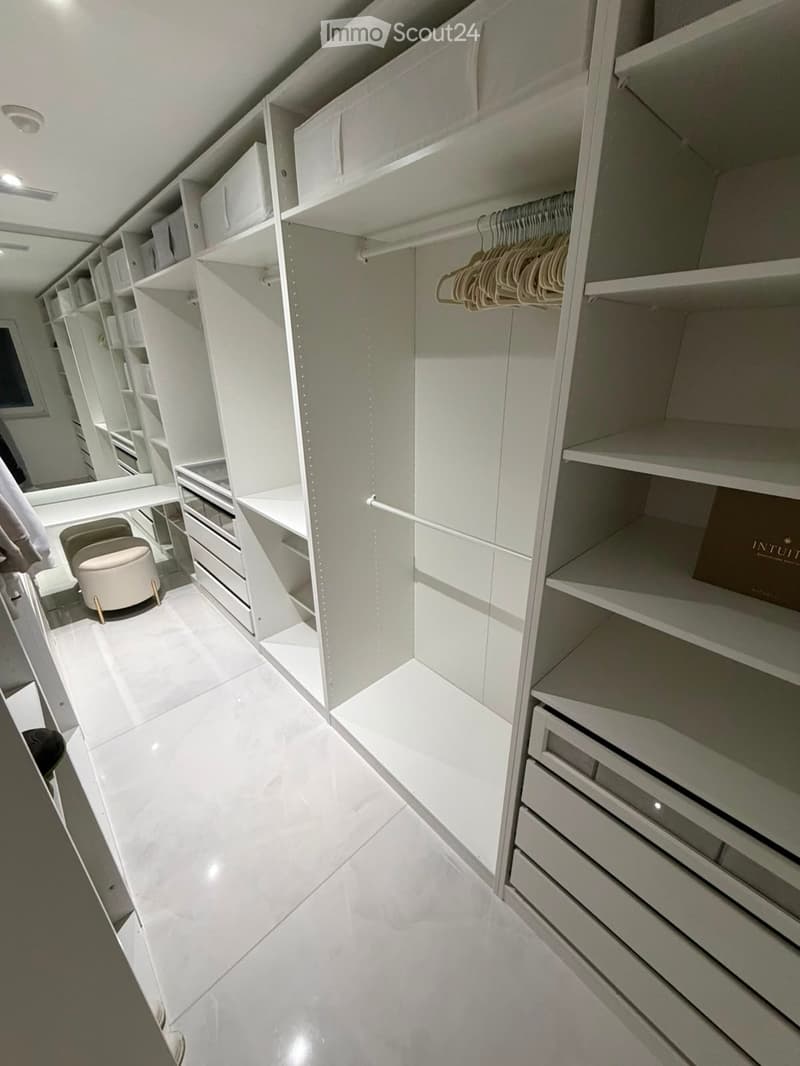 Walk-in closet with white built-in storage units, shelves, drawers, and a small beige stool.