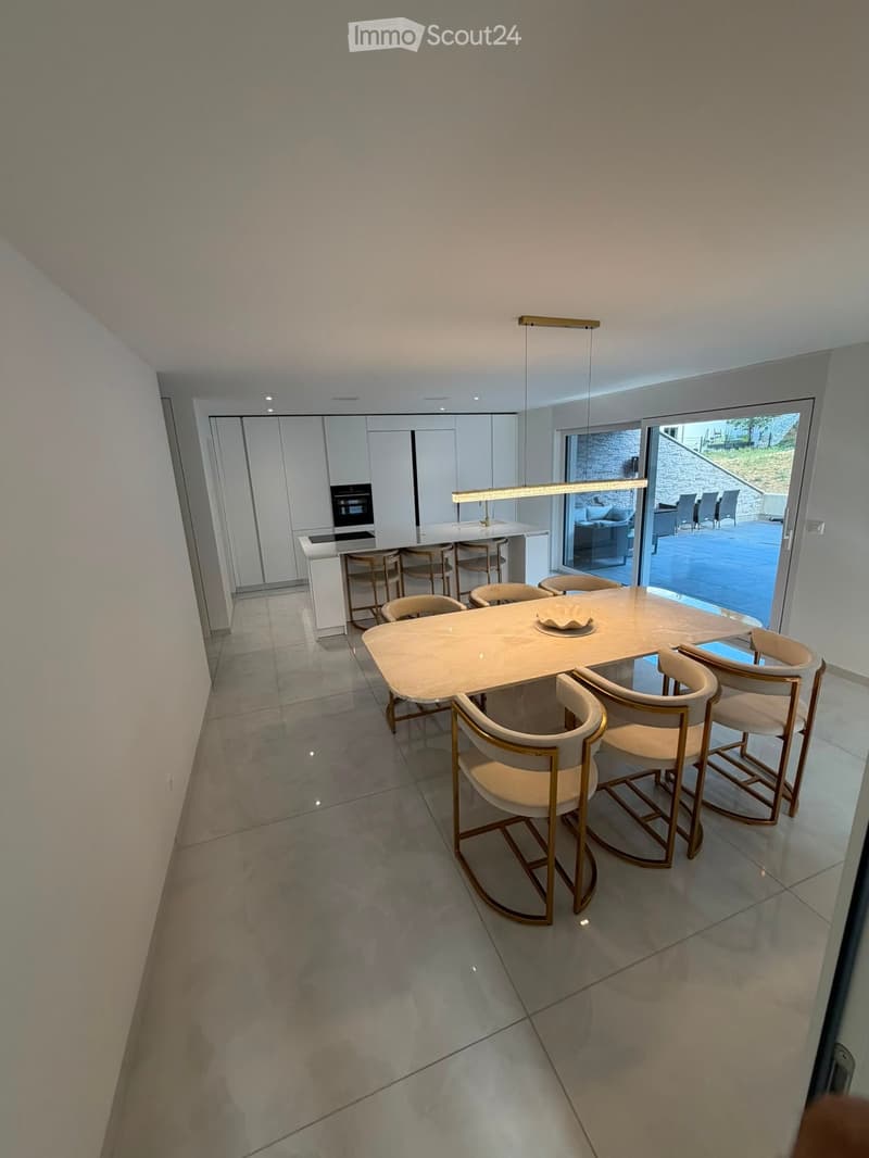 Modern dining area with a large white dining table surrounded by six beige chairs with gold accents, a kitchen island with bar stools, and built-in cabinets with an oven.