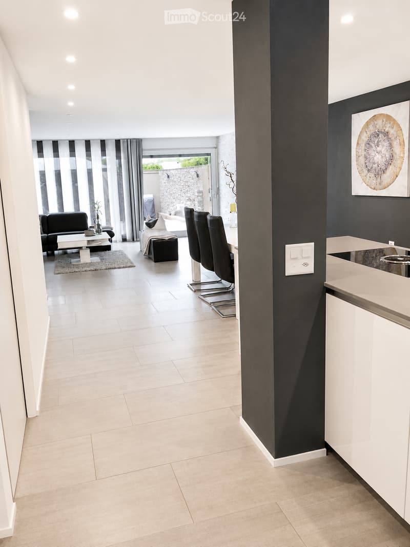 Modern open-plan living and dining area with light-colored tiles, a black accent wall, and contemporary furniture including a sofa, coffee table, dining chairs, and a small table with a lamp.