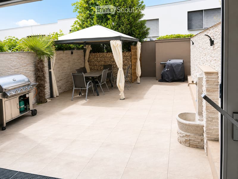 A modern outdoor patio with a tiled floor, a covered seating area with a table and chairs, a grill, a fire pit, and a covered barbecue area.