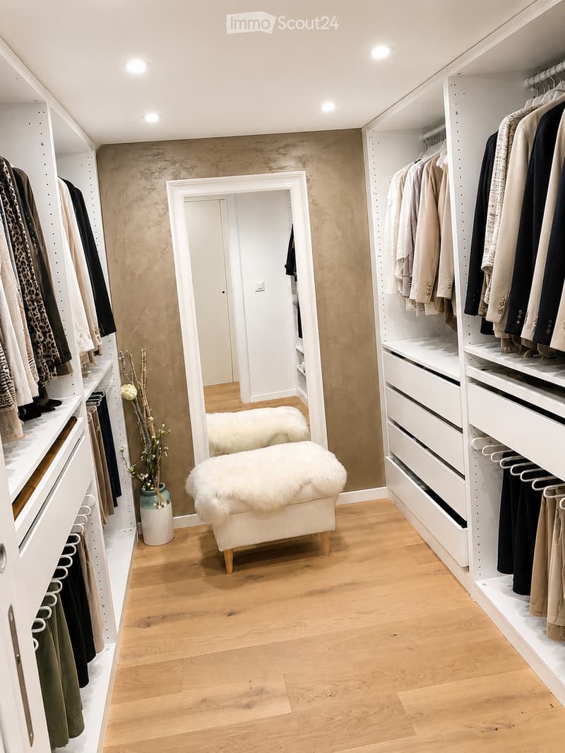 A modern walk-in closet with built-in white wardrobes, wooden flooring, a large mirror, a white fur-covered stool, and a decorative vase with dried branches.