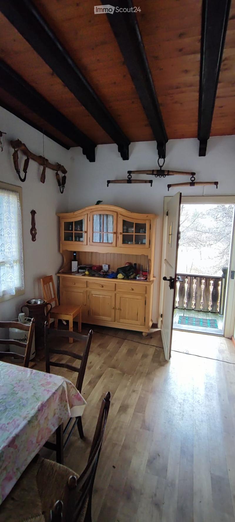 A cozy attic room with wooden furniture, including a table, chairs, and a wooden cabinet with glass doors. The room has exposed wooden beams on the ceiling and a door leading to a balcony.