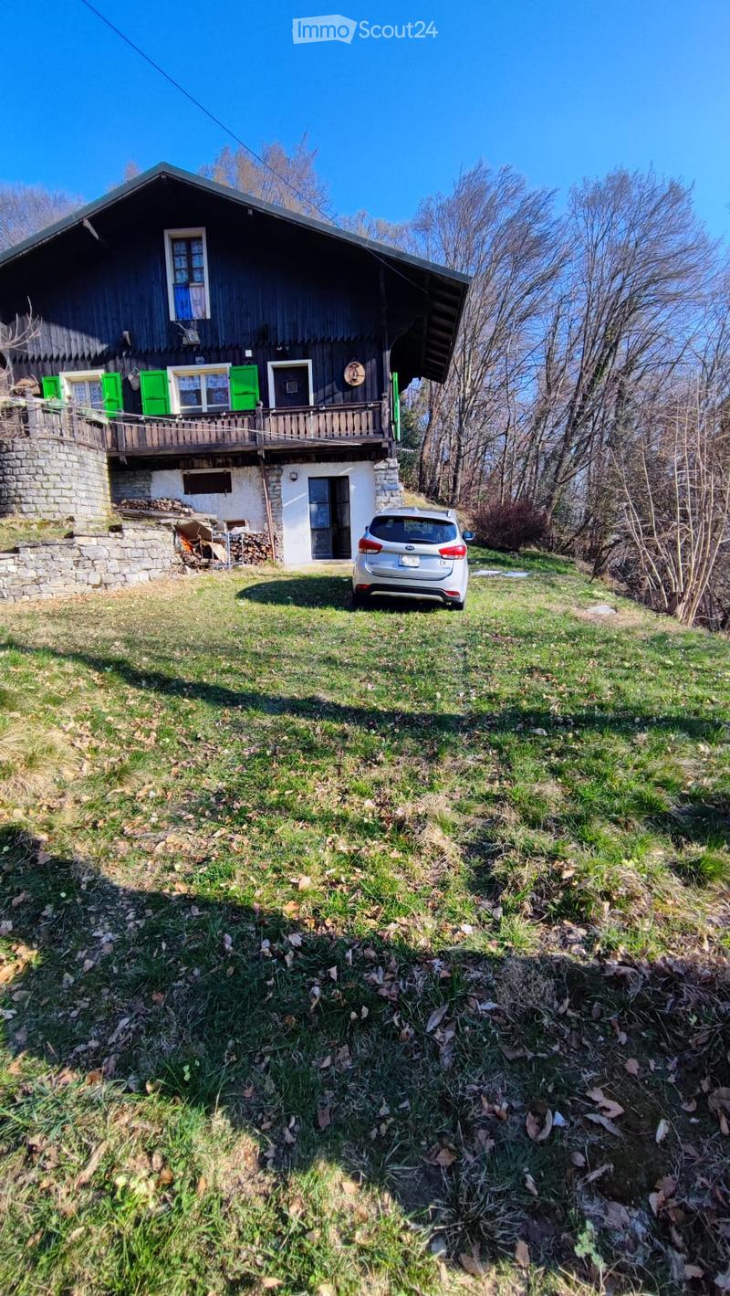 A two-story wooden house with green shutters and a stone base, featuring a balcony on the upper level and a car parked in front.