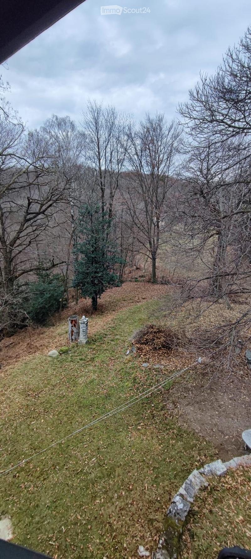 A grassy outdoor area with scattered trees, a stone pillar, and a view of a wooded landscape.