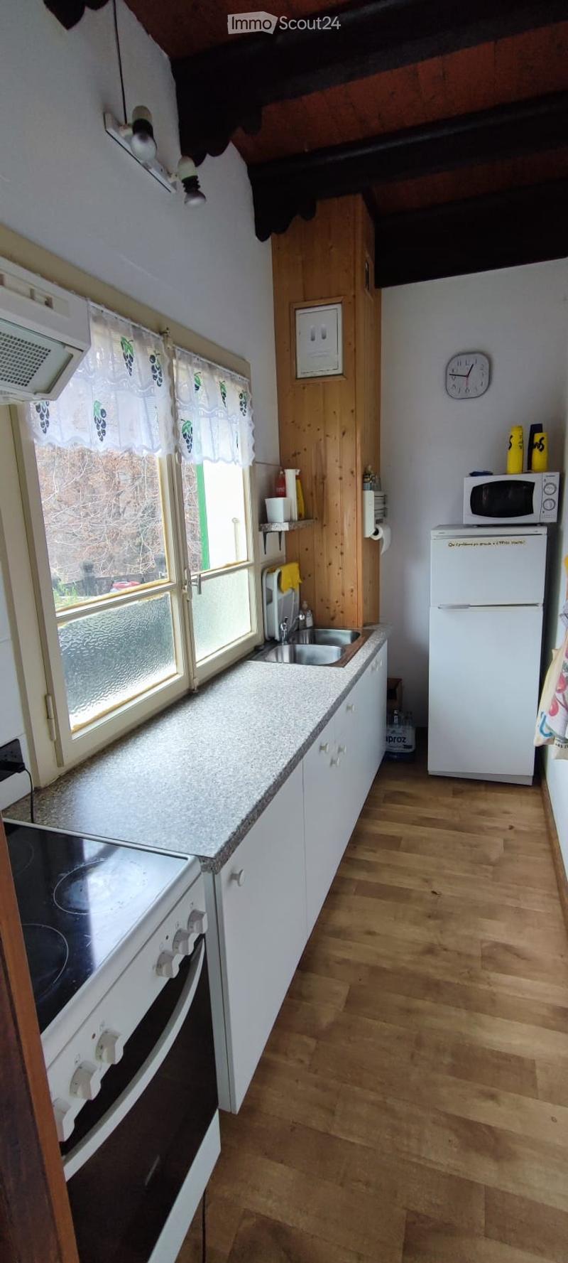 Bright kitchen with white cabinets, granite countertop, wooden floor, and stainless steel appliances including a refrigerator, microwave, and oven. A window with a curtain offers natural light.