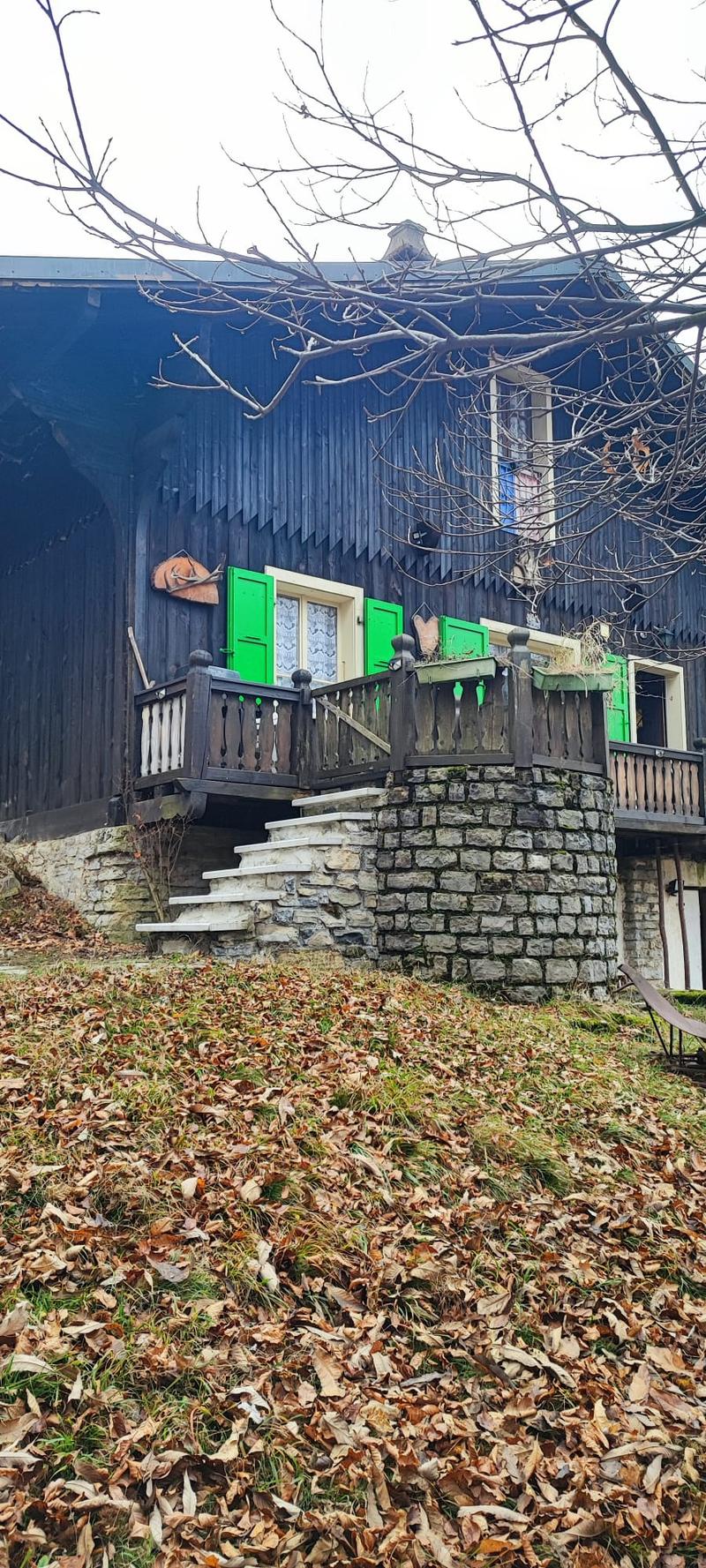 Two-story wooden house with green shutters, stone steps leading to a wooden deck, and a stone retaining wall covered in leaves.