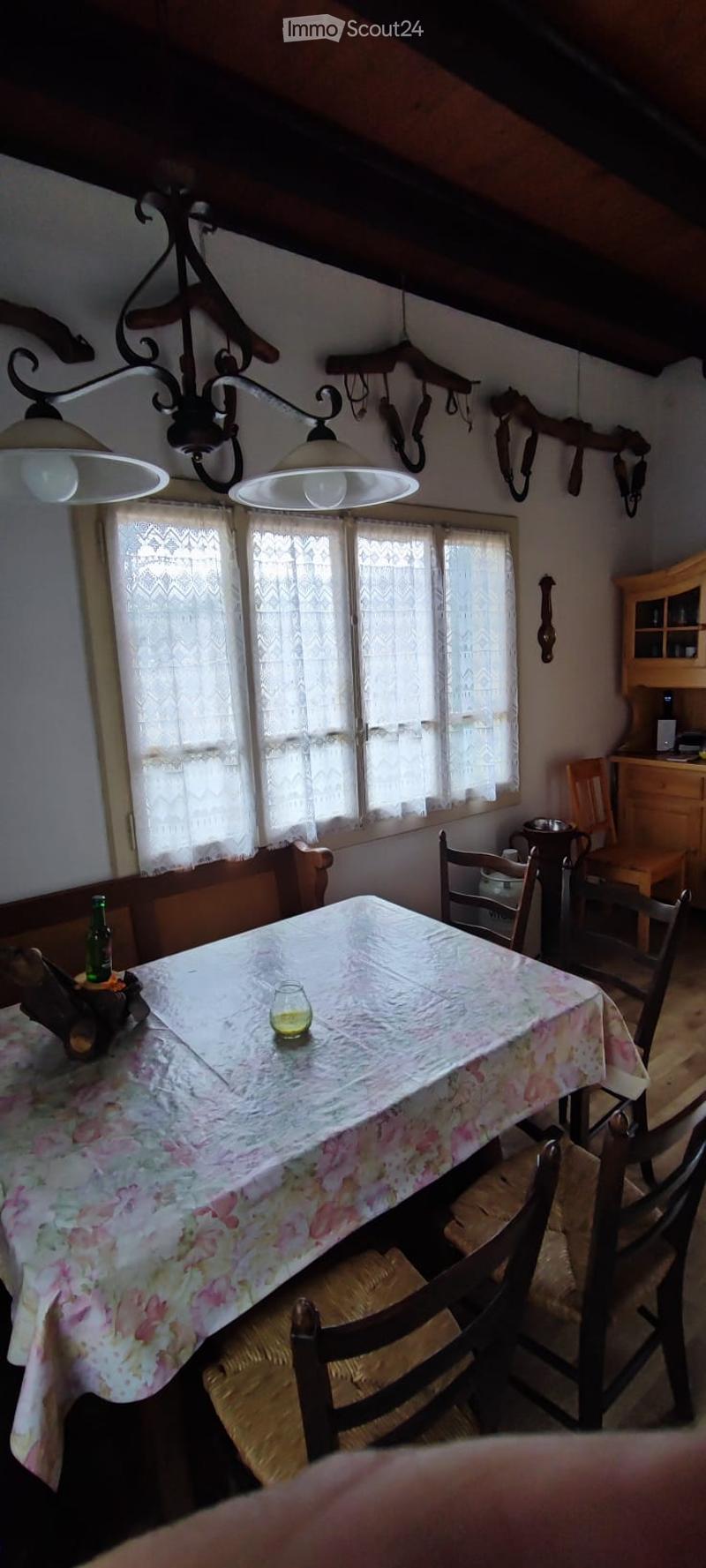 A cozy dining area with wooden chairs, a floral tablecloth, and a chandelier hanging from the ceiling. The room features wooden furniture, a window with lace curtains, and a small cabinet with glass doors.