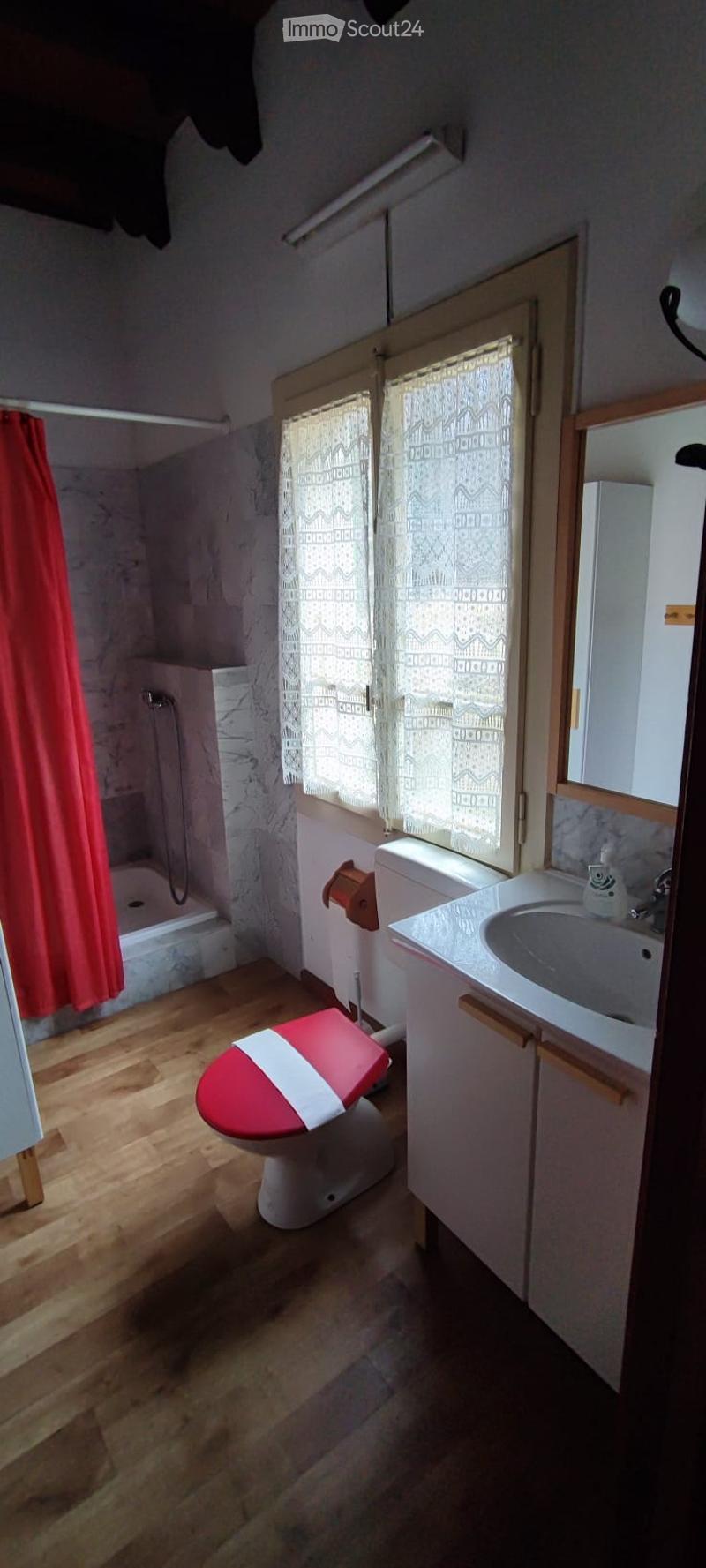 Bathroom with wooden flooring, a red toilet seat, a white sink with a cabinet, a bathtub, and a window with lace curtains.