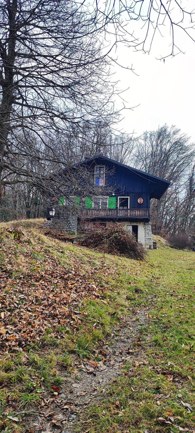 A two-story house with a dark blue exterior and green shutters, situated on a grassy slope with a stone foundation and a small balcony.