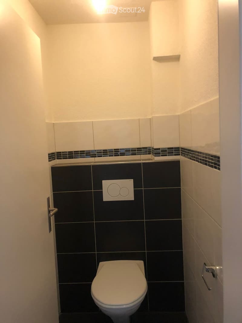 Modern bathroom with white walls, black tiles, and a toilet. Features a sleek design with a small shelf and a light switch.