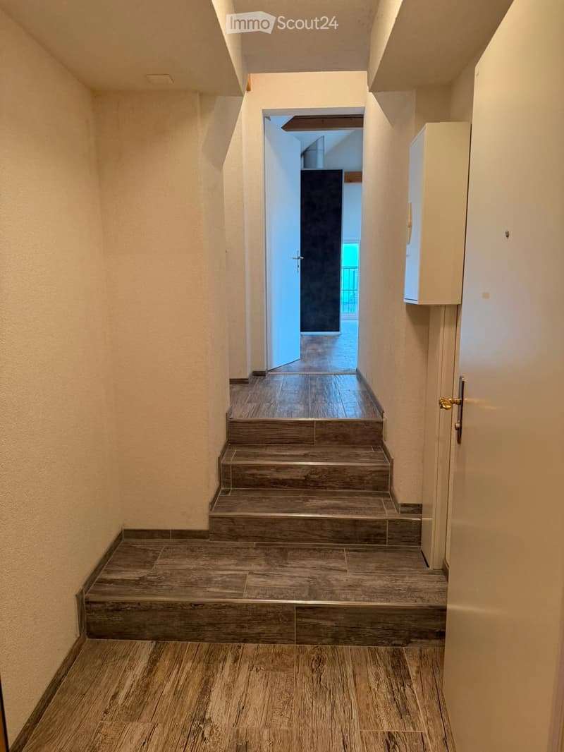 A narrow hallway with wooden flooring and a staircase leading upwards, featuring white walls and a closed door on the right side.