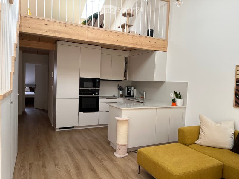 Modern, open-plan living and kitchen area with a yellow sofa, cat tree, white kitchen cabinets, microwave, oven, sink, and a small potted plant on the counter.
