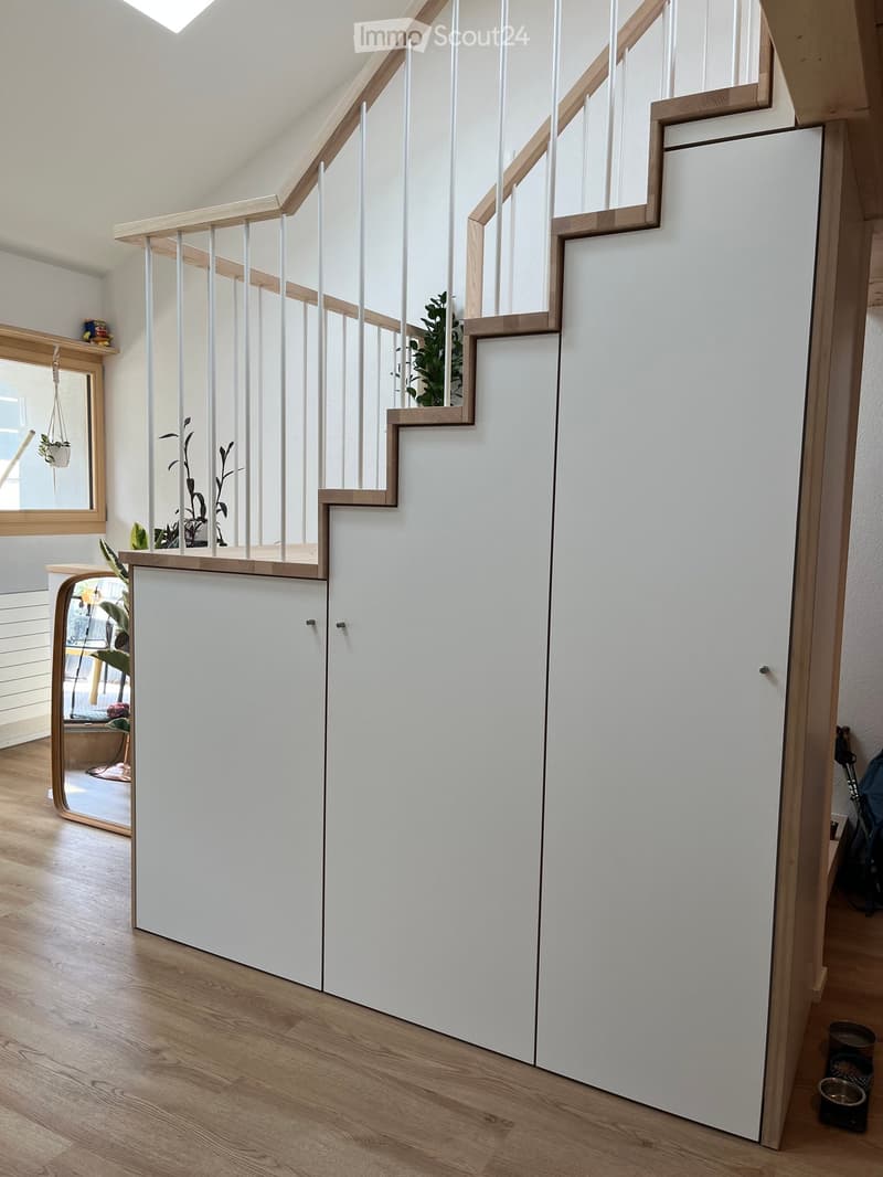 Modern hallway with a staircase featuring white railings and wooden steps, white walls, and a wooden floor. There are small potted plants and a mirror visible in the background.