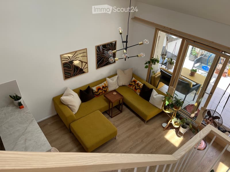 A modern living room with a green sectional sofa, wooden coffee table, side tables, potted plants, and a fan. The room features large windows with a view of a balcony area with additional seating and plants.