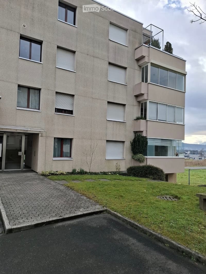 Modern multi-story building with large windows, balconies, and a landscaped lawn area.