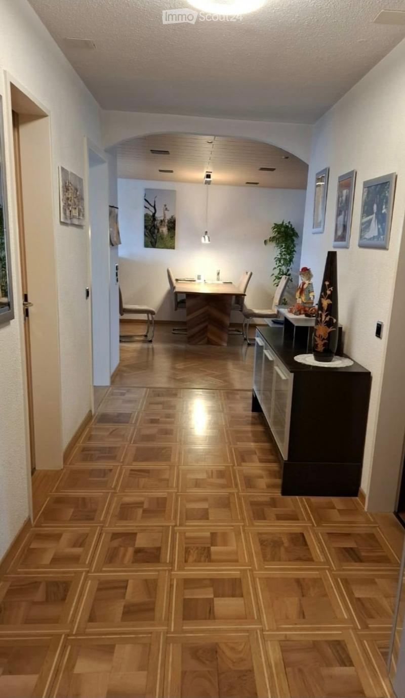 A well-lit hallway with wooden flooring, leading to a small dining area with a table and chairs, a cabinet with a vase, and framed pictures on the walls.