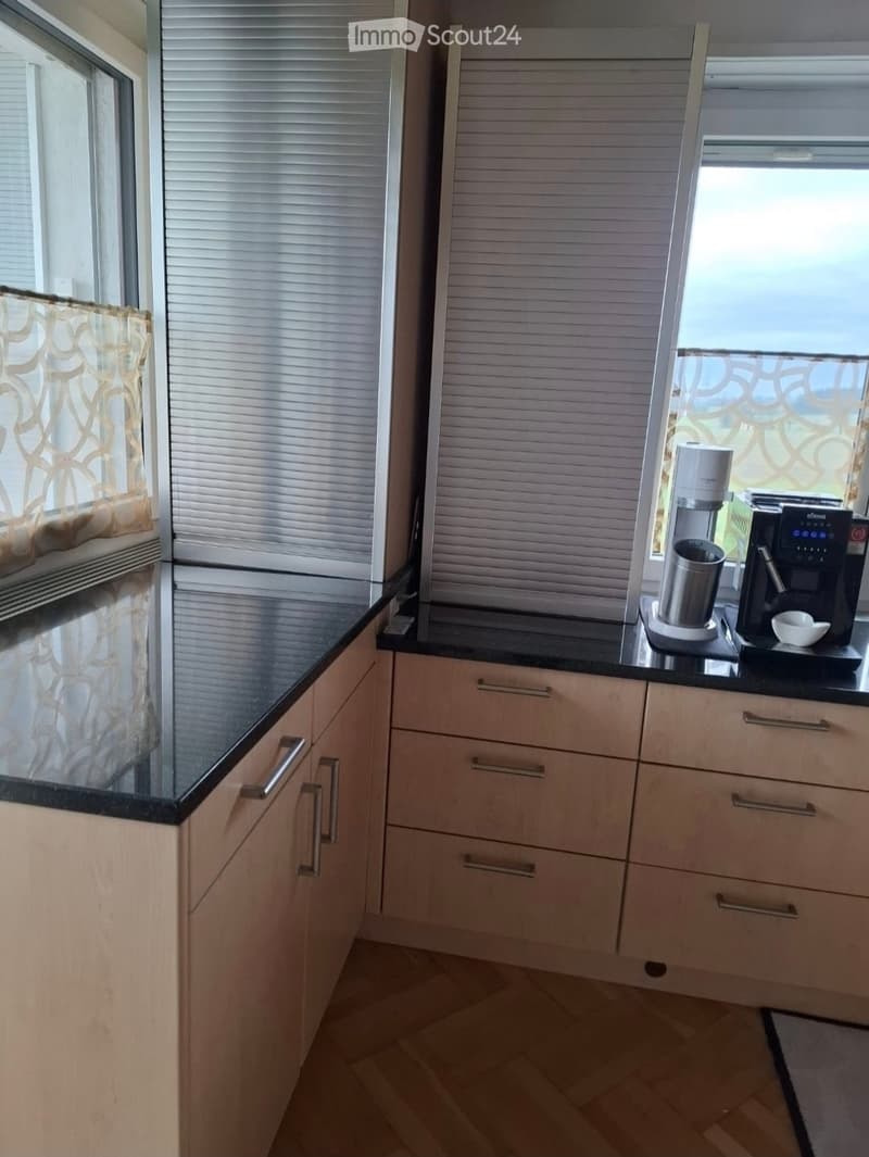 Modern kitchen with light wood cabinets, black countertops, and a coffee maker on the counter. The room features large windows with blinds and a view of the outdoors.