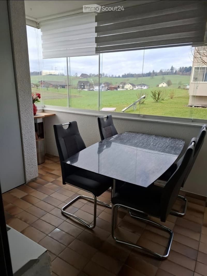 Dining area with a glass table, four black chairs, and a small flower vase on a side table near a large window with blinds, overlooking a green landscape.