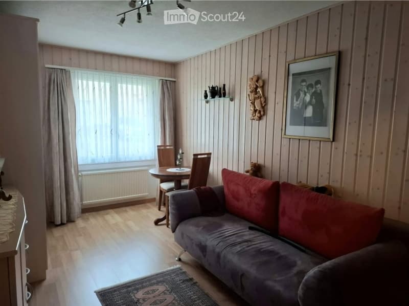 A cozy living room with wooden walls, a grey sofa with red cushions, a small dining area with a round table and chairs, a shelf with decorative items, and a framed picture on the wall.