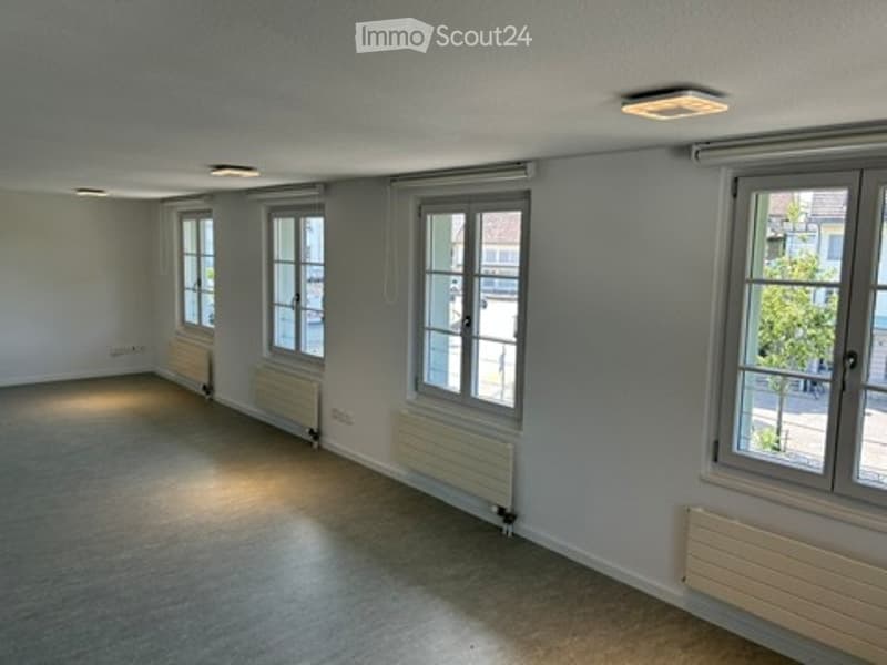 Empty room with light grey walls, carpeted floor, and multiple windows with white frames. Radiators are visible under each window, and the ceiling has recessed lighting.