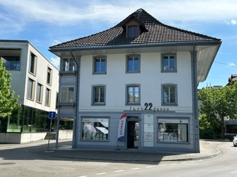 A three-story building with a classic European architectural style, featuring a tiled roof, multiple windows, and a storefront with signage reading "farm2kasse" and a RE/MAX logo.