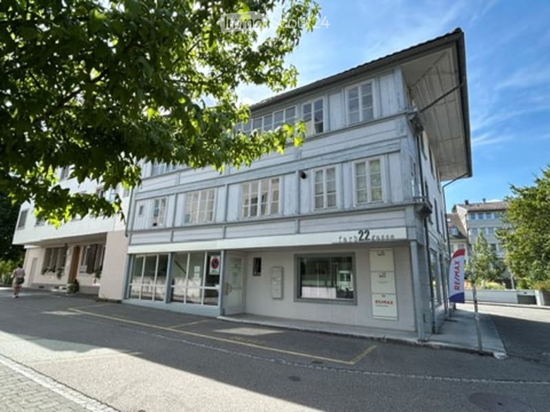 Two-story building with white facade, multiple windows, and a storefront on the ground floor. The building has a modern design with a flat roof and is surrounded by trees.