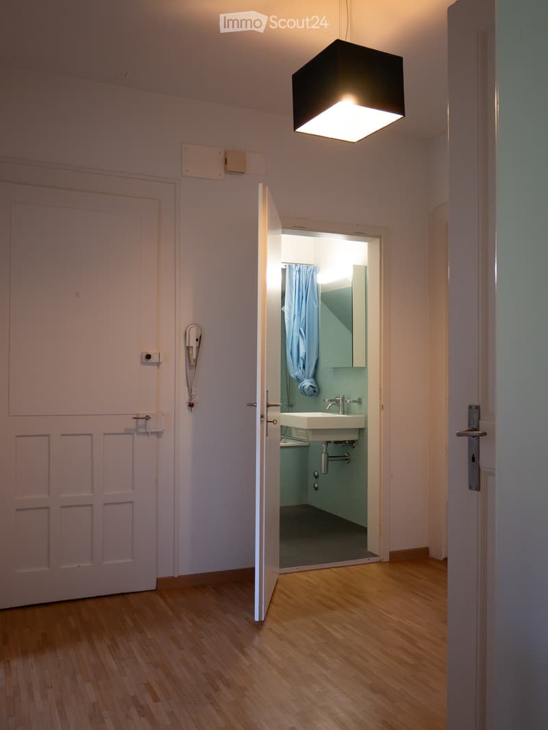 A narrow hallway with wooden flooring and white walls, leading to an open door that reveals a small bathroom with a sink, mirror, and light blue tiles.