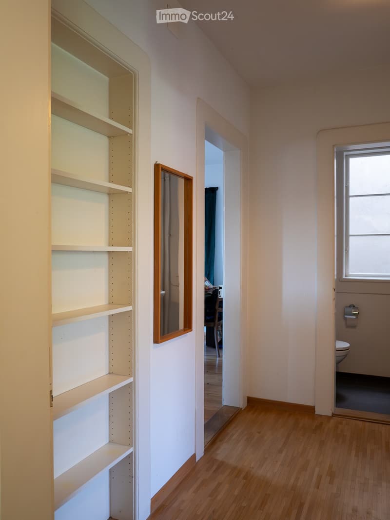 Empty hallway with wooden flooring, white walls, built-in shelves, and a small window. A partially open door leads to a room with a visible toilet and a mirror.