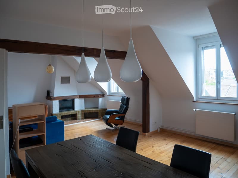 Bright attic room with wooden floors, exposed wooden beams, large windows, fireplace, dining table, chairs, and a comfortable sofa.