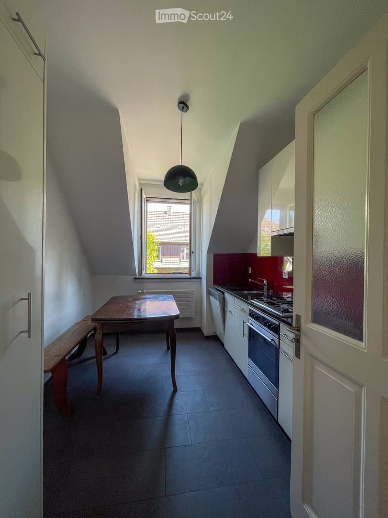 Compact kitchen with a wooden table and bench, white cabinets, stainless steel oven, and a window offering a view of the roof.