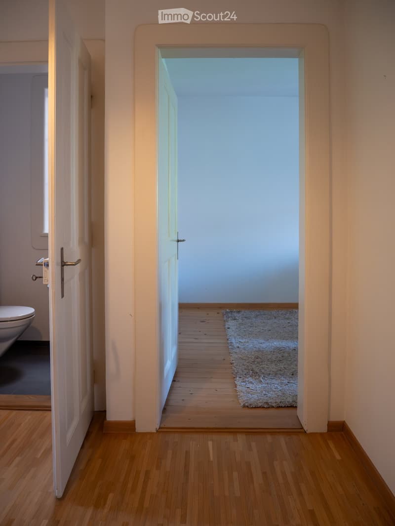 A narrow hallway with wooden flooring, light-colored walls, and a small white rug placed on the floor. An open door leads to another room with a visible toilet.