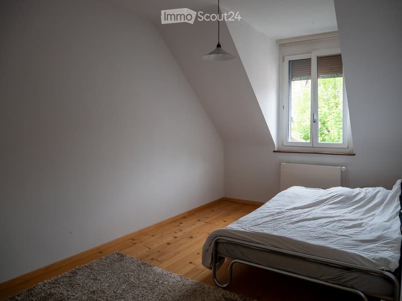 Spacious attic bedroom with wooden flooring, a single bed, a grey rug, and large windows with blinds. The room features white walls and a simple hanging light fixture.