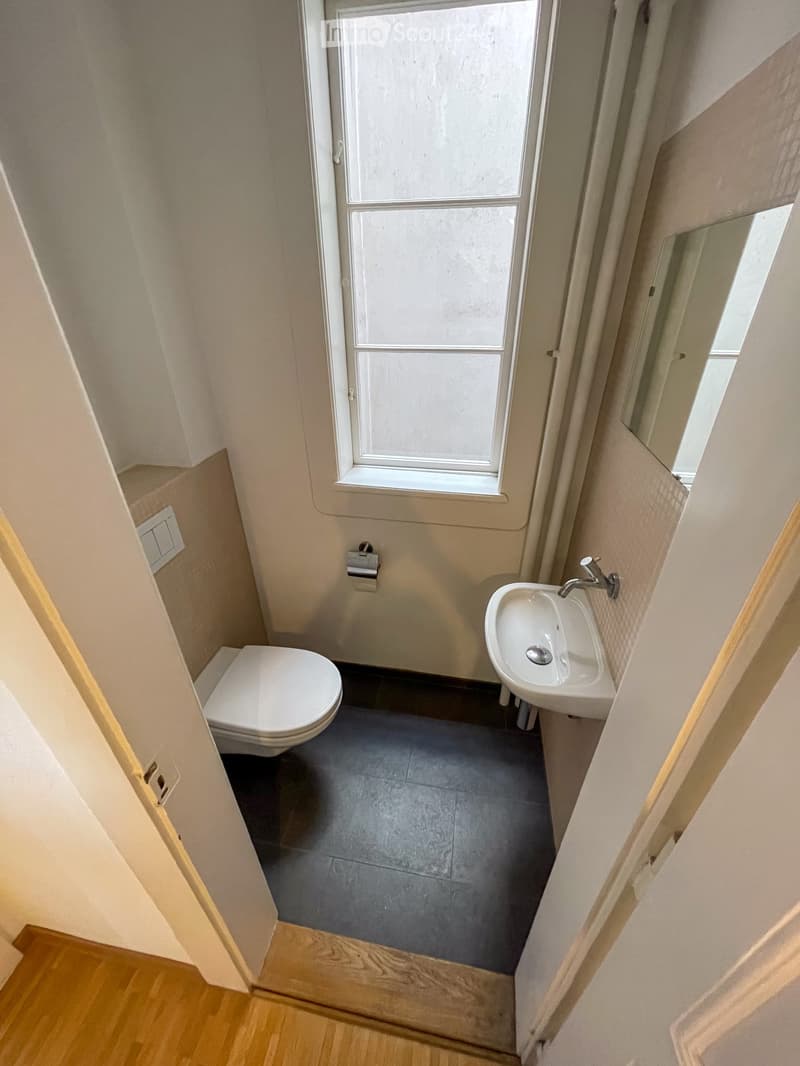 Compact bathroom with a toilet, sink, and window. Light-colored walls and dark tiled floor.