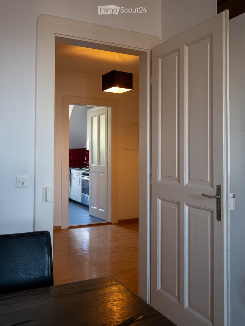 A hallway with wooden flooring leads to a kitchen area visible in the background. A black leather chair and a wooden table are in the foreground, with a modern light fixture hanging from the ceiling.