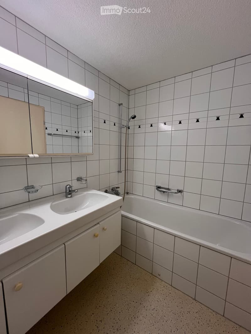 Bathroom with white tiled walls and floor, double sink with cabinets, bathtub, mirror with built-in lighting, and a showerhead.