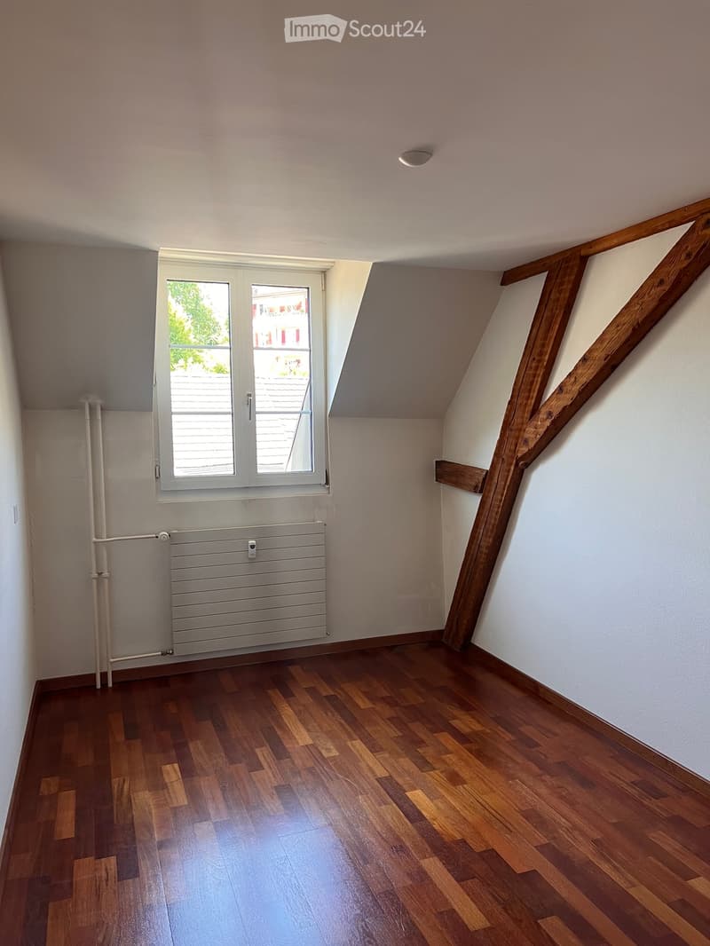 Empty attic room with wooden flooring, white walls, exposed wooden beams, and a window with blinds.