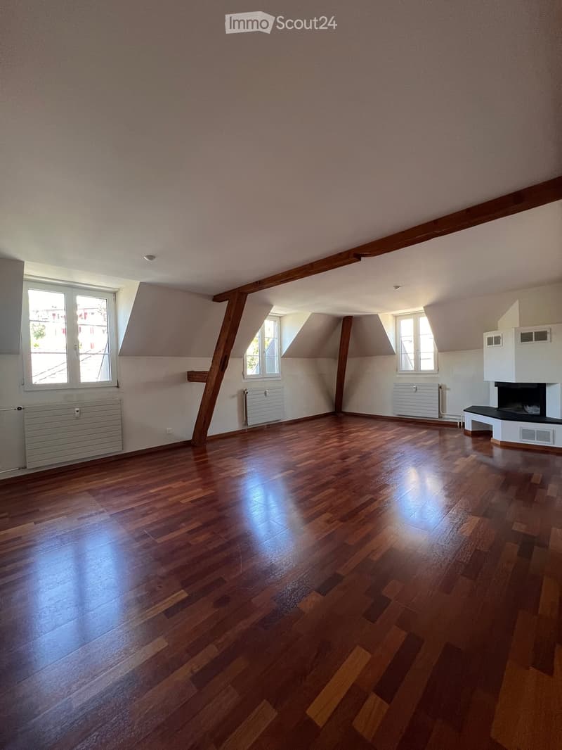 Empty room with wooden flooring, exposed wooden beams, and a fireplace. Natural light from multiple windows illuminates the space.